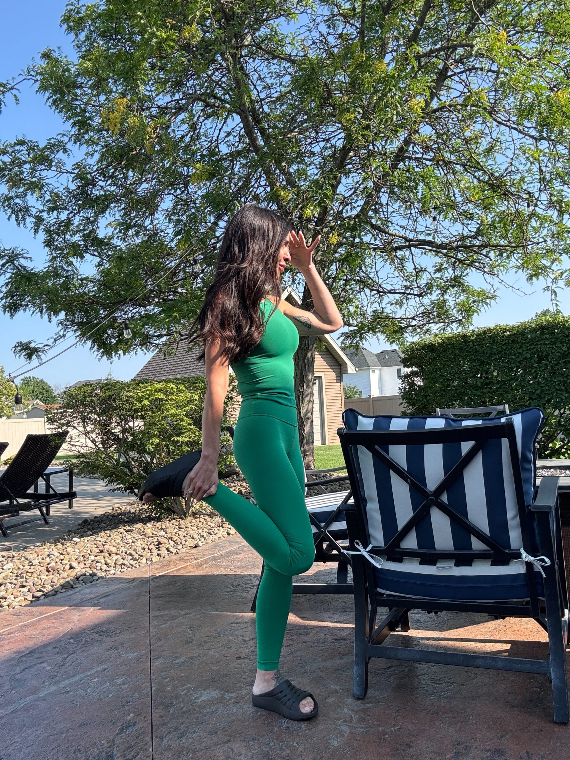 Woman in a green workout set stretching outdoors by a pool, wearing black recovery slides on a sunny day.